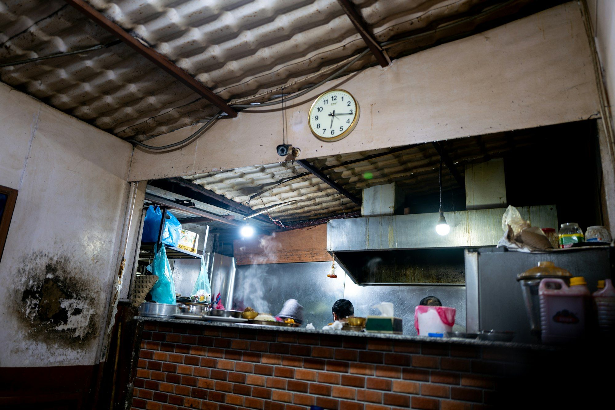 Dapur warung malam hari dengan atap seng, jam dinding bundar, konter bata merah, dan hood stainless di belakangnya