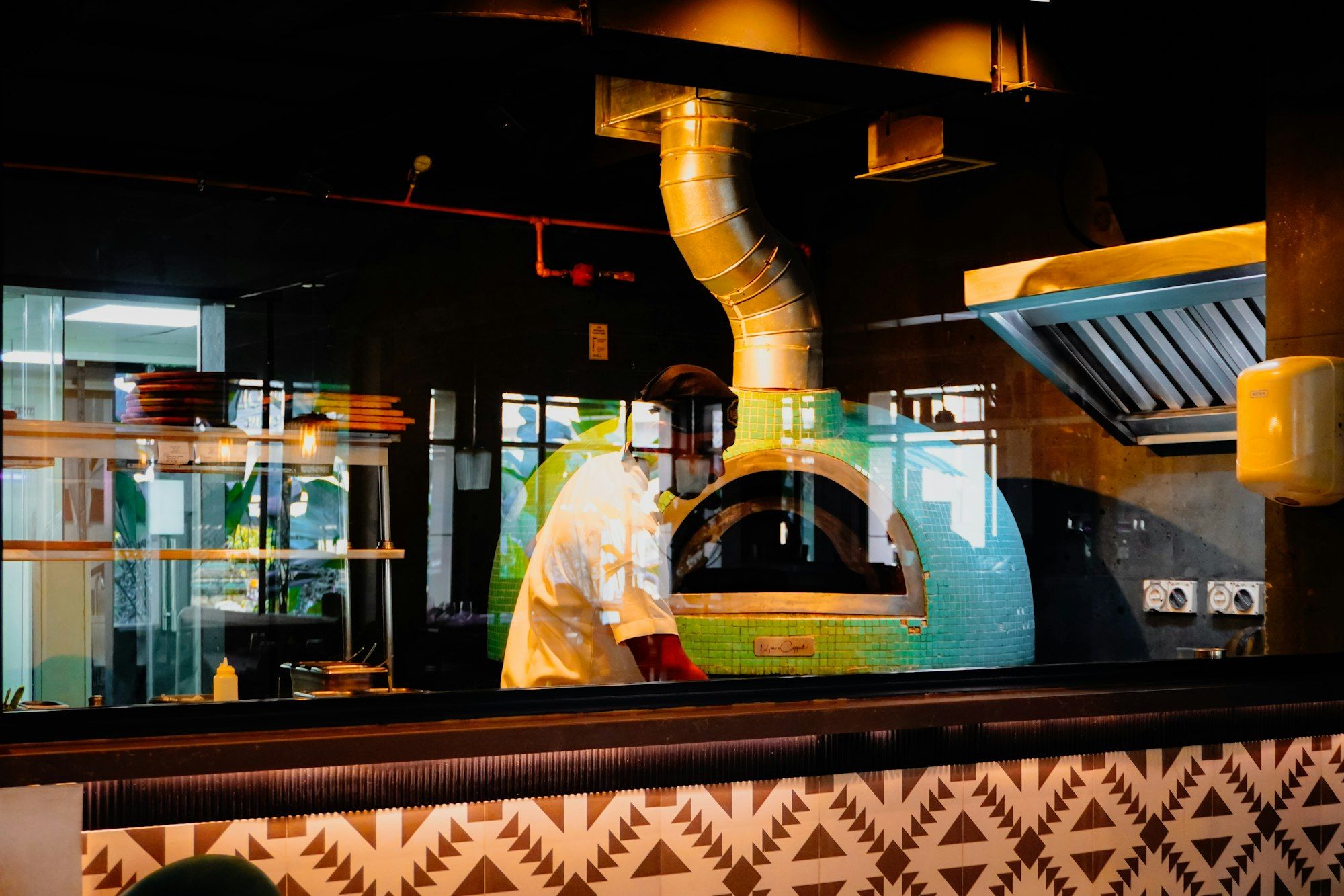 Dilihat dari balik kaca, juru masak berjaket putih berdiri di depan oven kubah berubin hijau toska dengan cerobong kuningan