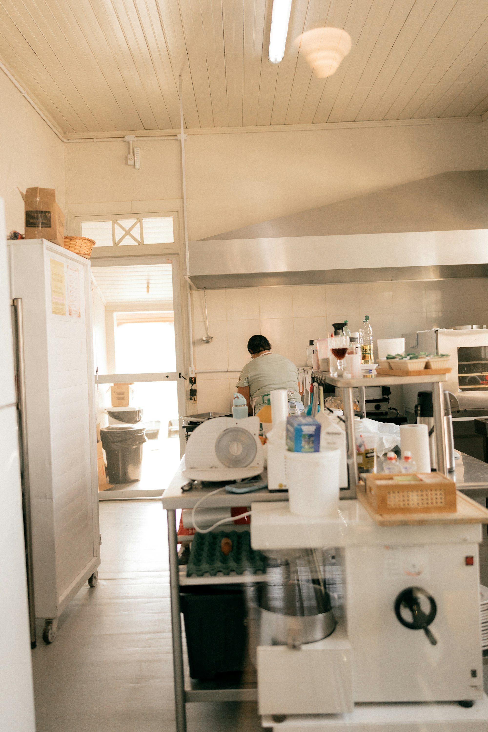 Dapur produksi serba putih, satu orang berkaus hijau muda bekerja membelakangi kamera di meja stainless, dengan mixer besar dan rak telur di depan