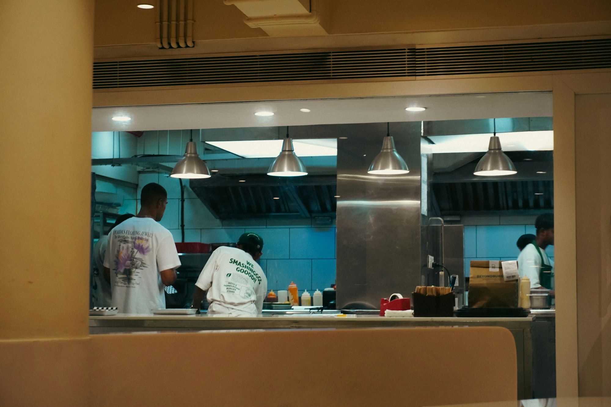 Dapur terbuka dilihat dari jendela saji: dinding ubin biru, lampu gantung baja, tiga orang berkaus putih bekerja di lini