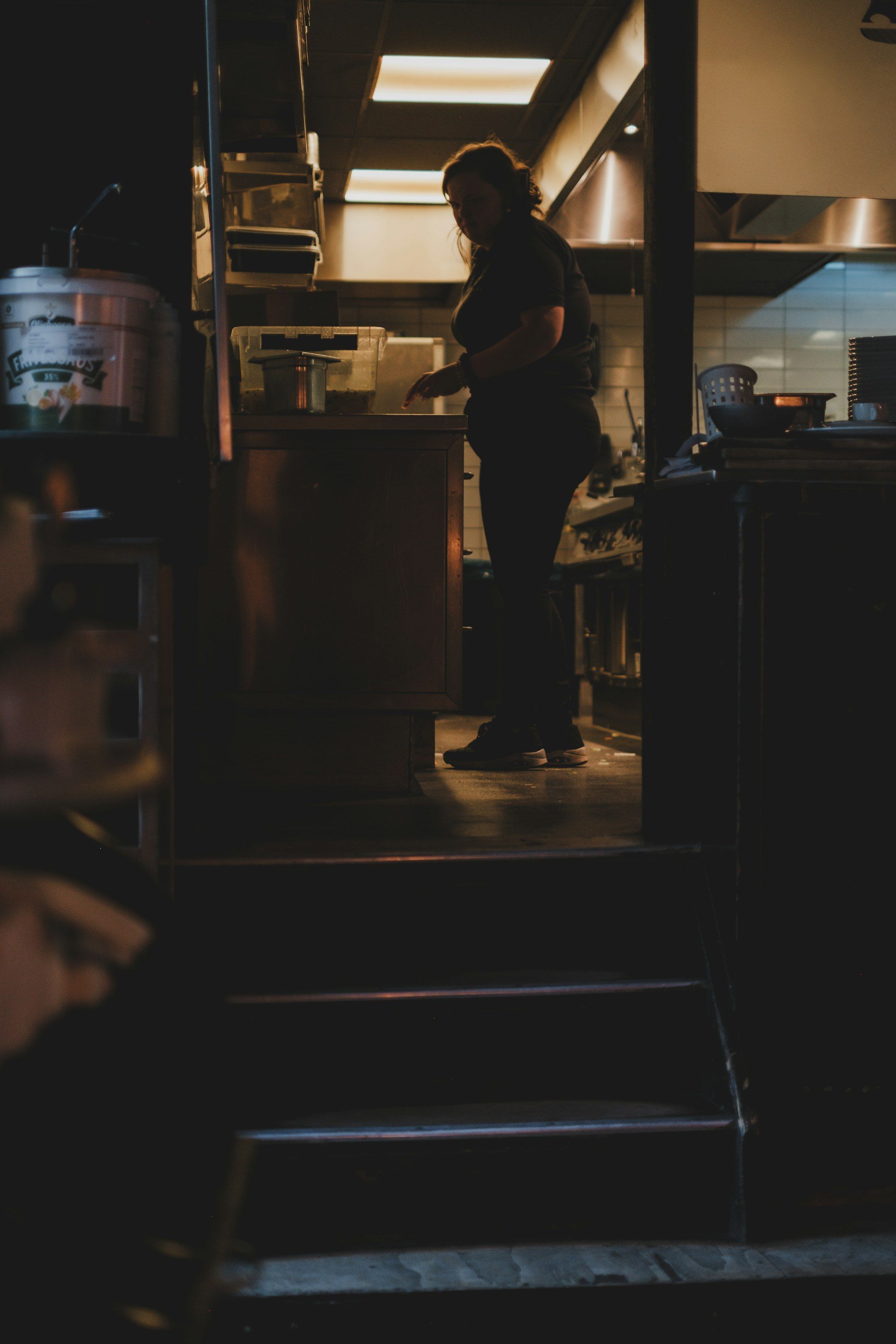 Dapur remang menjelang tutup, seorang perempuan berbaju hitam berdiri di meja persiapan di bawah lampu neon, dinding ubin putih di belakangnya