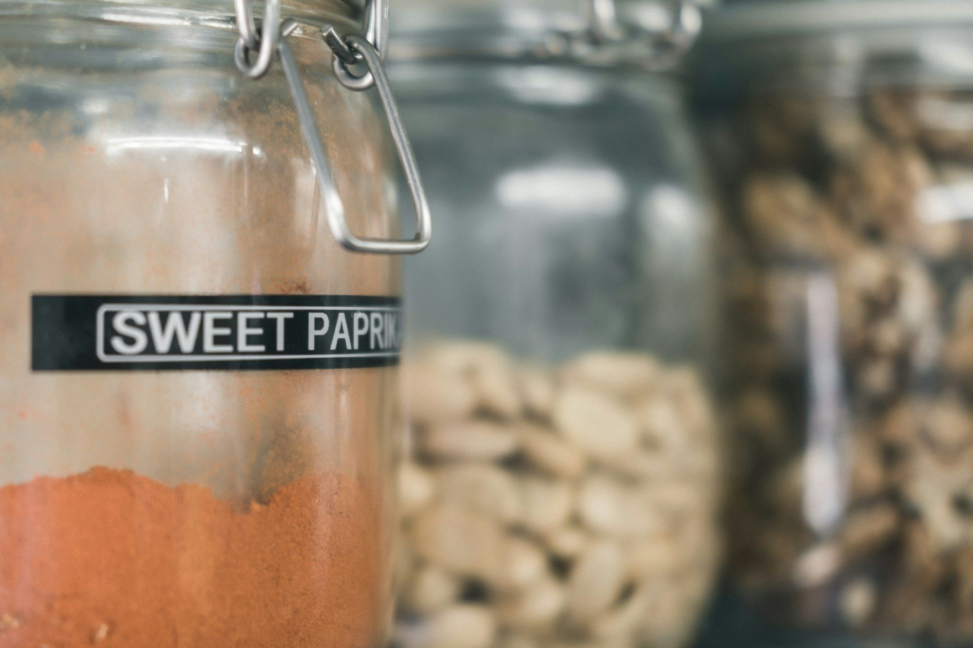 Toples kaca berlabel SWEET PAPRIKA berisi bubuk paprika merah, dua toples bumbu lain di belakangnya