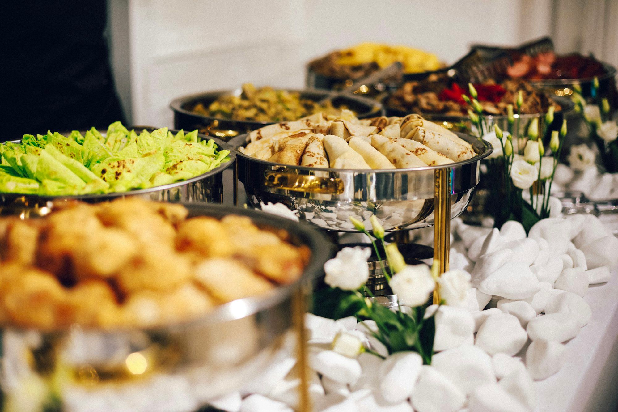 Meja prasmanan dengan chafing dish berisi dadar gulung, selada, dan gorengan, dihias bunga putih dan batu putih