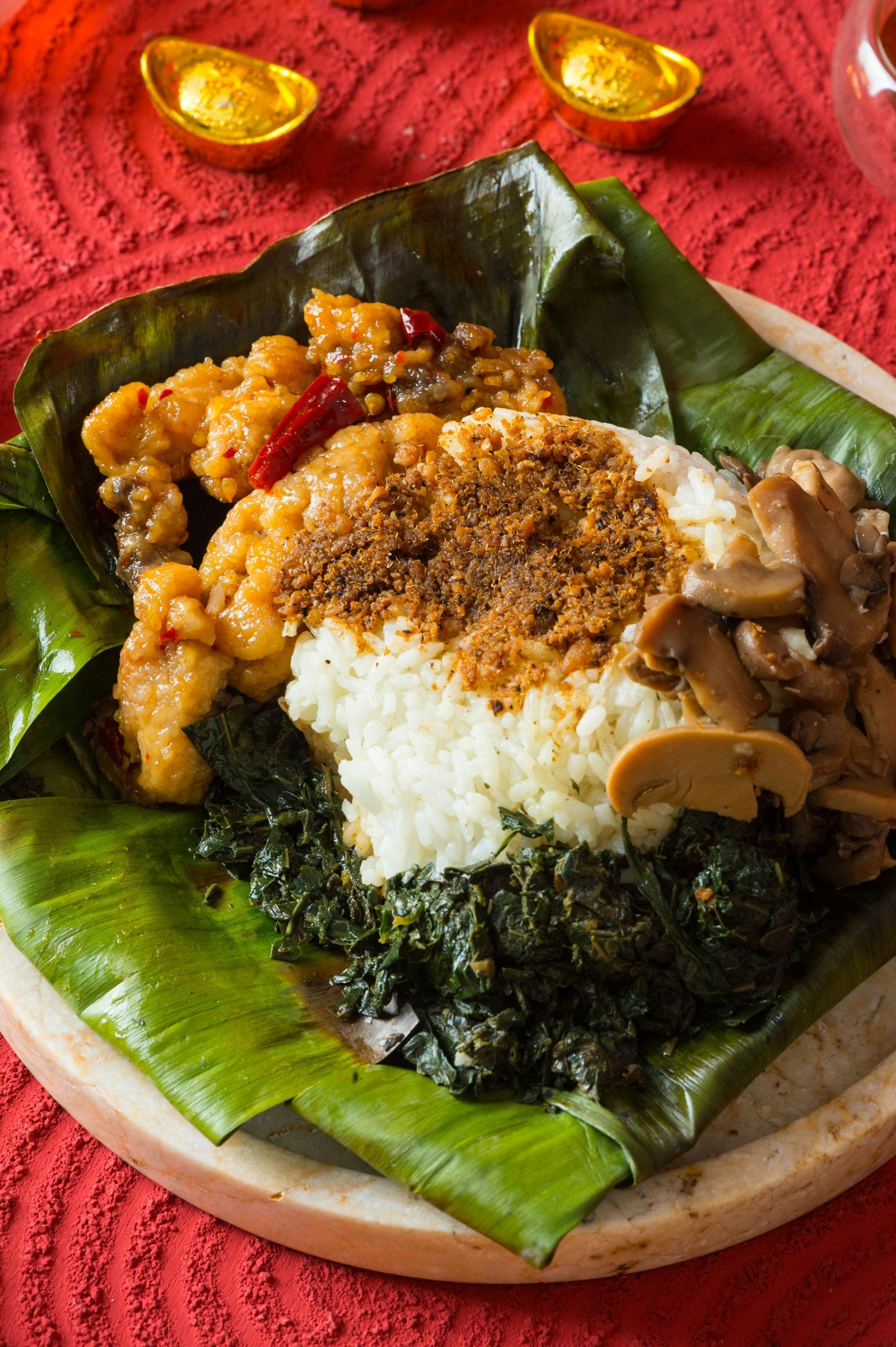 Satu porsi nasi putih di atas daun pisang dengan taburan serundeng, ayam goreng saus manis, tumis daun singkong, dan jamur