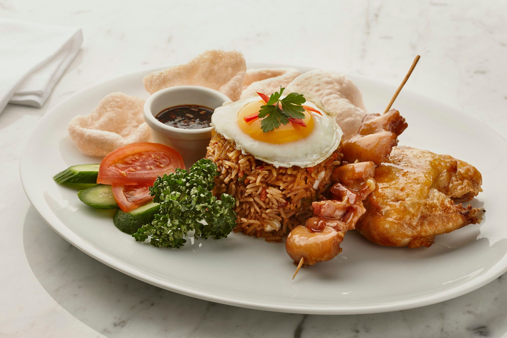 Piring putih berisi nasi goreng dengan telur mata sapi di atasnya, satu tusuk sate ayam, ayam goreng, kerupuk, timun, dan tomat
