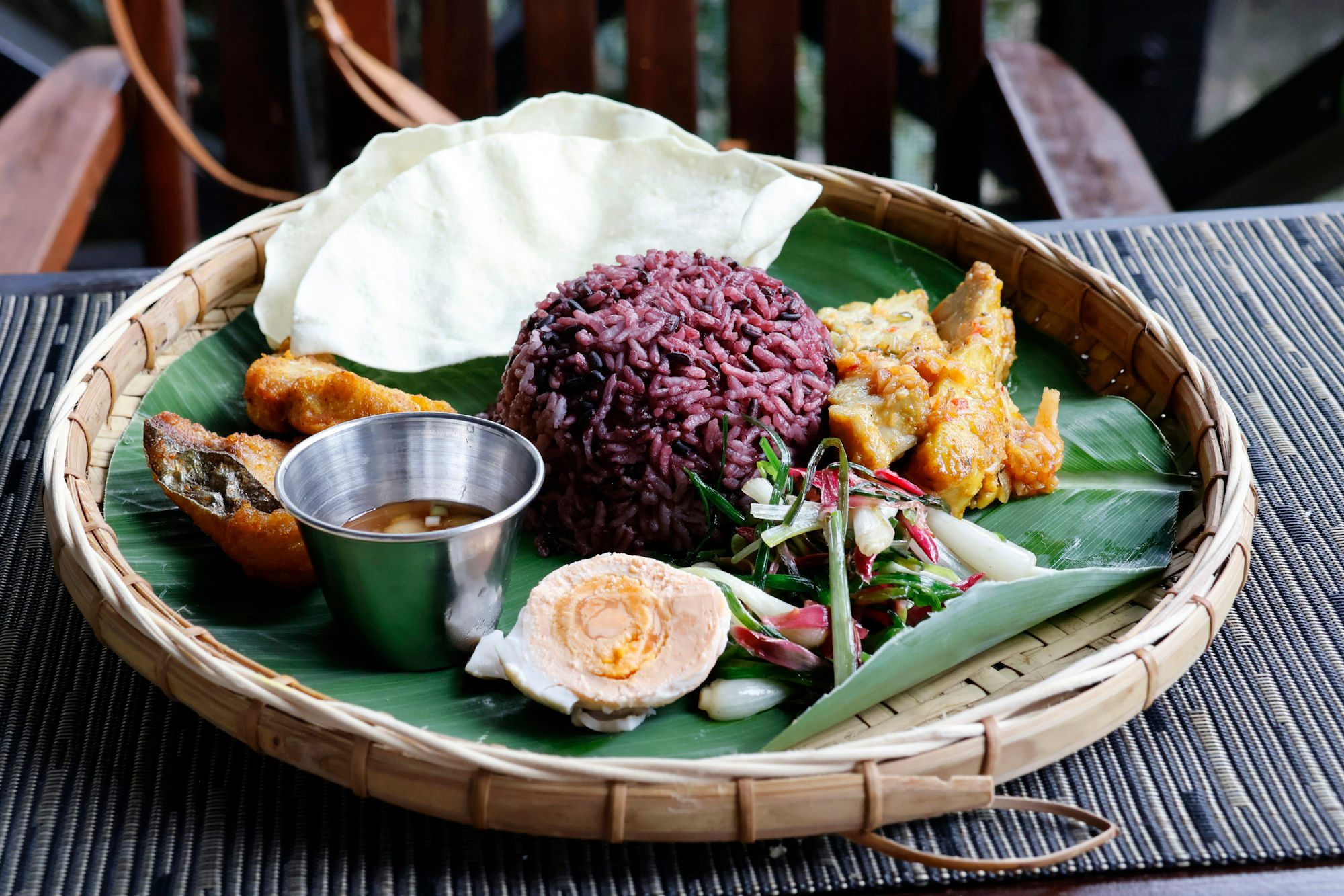 Nampan bambu beralas daun pisang berisi nasi hitam, tempe bumbu kuning, ikan goreng, telur asin, tumis kacang panjang, dan kerupuk