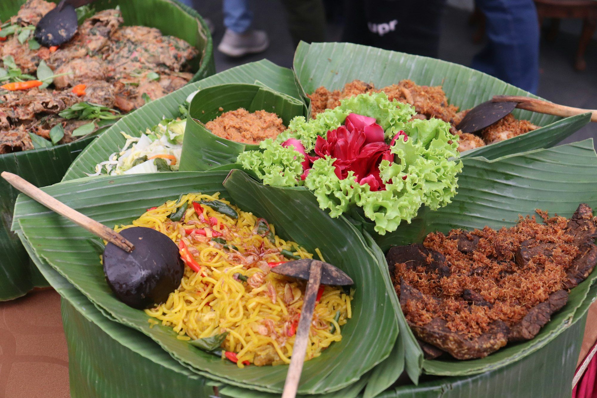 Nampan besar beralas daun pisang berisi mi goreng kuning, daging berselimut serundeng, sambal dalam wadah daun, dan acar, dihias selada keriting dan bunga merah