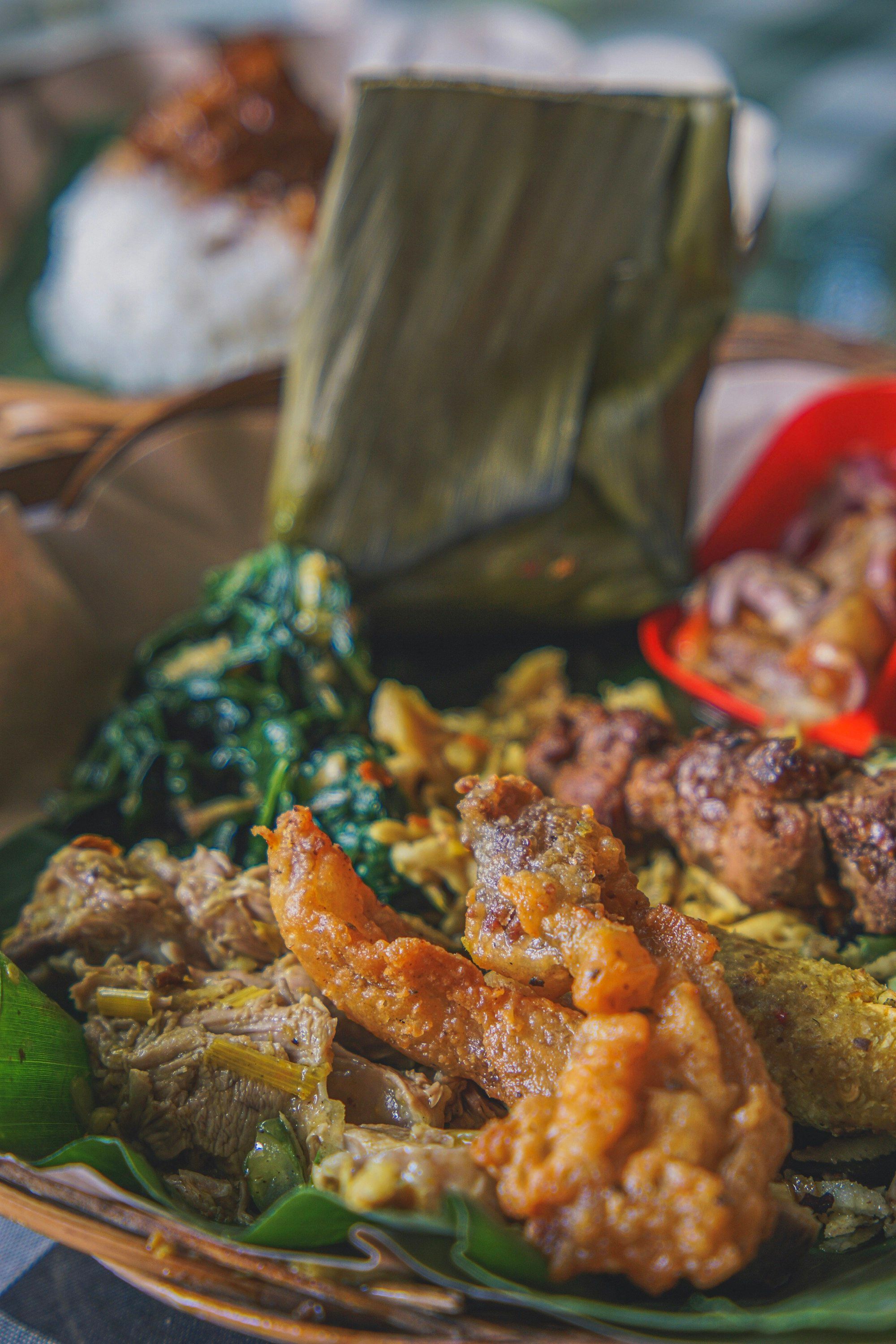 Piring nasi campur Bali dari dekat: ayam suwir, tempe goreng, urab kacang panjang, sate lilit, dan gorengan di atas daun pisang