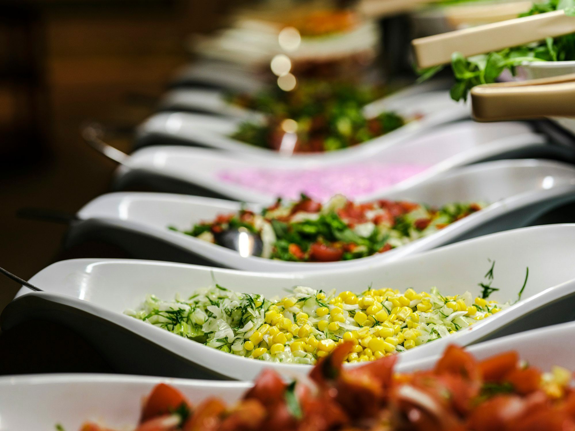 Deretan mangkuk saji putih melengkung berisi salad kol jagung, salad tomat, dan sayur hijau di meja prasmanan